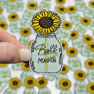 May include: A green mason jar with a yellow sunflower inside. The jar has the text "Ball's Mason" on the front.
