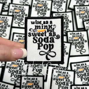 May include: A white fabric patch with black embroidered text that reads "wild as a mink, but sweet as soda pop". The patch has a black border and a small infinity symbol at the bottom.
