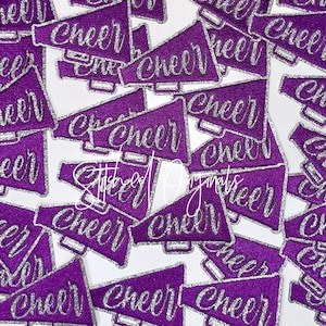 May include: A collection of purple glitter megaphone-shaped patches with the word "Cheer" in silver script. The patches are scattered on a white background, creating a dynamic and festive display.