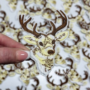 May include: A close-up of a white embroidered iron-on patch of a brown and yellow deer head with antlers.