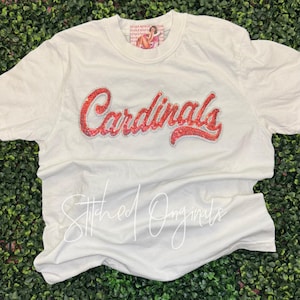 May include: A cream-colored short-sleeve t-shirt featuring the word Cardinals in red script lettering across the chest. The text is embellished with small red rhinestones. A small rectangular tag with a graphic illustration is visible at the neckline.