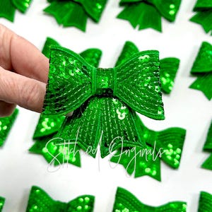 May include: A close-up of a bright green sequined bow held by a hand, with several similar bows in the background. The bows are made of shiny, emerald green sequins and have a classic bow shape. The text "Stitched Originals" is visible.