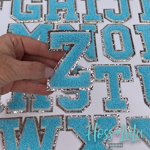 May include: A light blue chenille letter "Z" patch with a silver glitter border, held by a hand. Other chenille letter patches are visible in the background. The patches are for apparel or craft projects.
