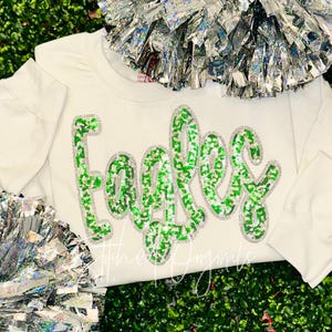 May include: Cream-colored sweatshirt with the word "Eagles" in large, green sequined letters. The letters are outlined in silver sequins. Silver pom-poms are visible in the background. The text "Stitched Originals" is at the bottom.