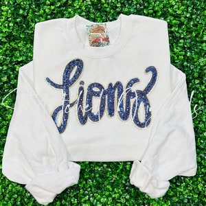 May include: A white sweatshirt with long sleeves. The word "fierce" is written across the front in a cursive font, with blue sequins and a gold outline. A small tag is visible near the neckline.