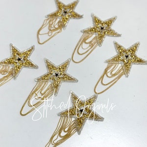 May include: Five gold star-shaped embellishments with a black center and a chain fringe. The embellishments are on a white background.