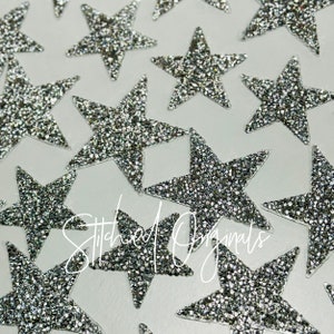 May include: A close-up of a collection of silver glitter star-shaped embellishments. The stars are scattered on a white surface. The text "Stitched Originals" is visible in the lower center of the image.