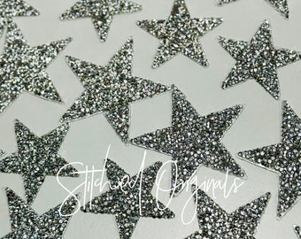 May include: A close-up of a collection of silver glitter star-shaped embellishments. The stars are scattered on a white surface. The text "Stitched Originals" is visible in the lower center of the image.