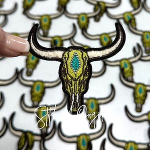 May include: A close-up of a green embroidered bull skull patch with a turquoise gem in the center. The patch is surrounded by other similar patches.