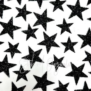 May include: A sheet of black glitter star stickers. The stars are various sizes and are arranged in a random pattern on a white background.