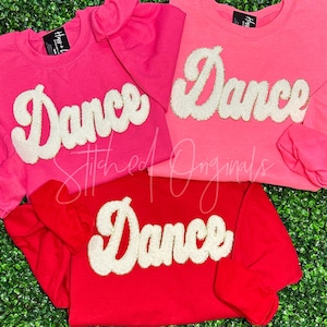 May include: Three sweatshirts with the word "Dance" in white glitter letters on a pink and red background. The sweatshirts are all long-sleeved and have a crew neck.