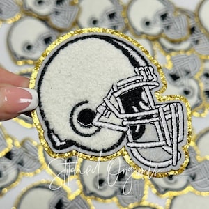 May include: A white and black embroidered football helmet patch with a gold glitter border. The patch is made of felt and has a stitched design.