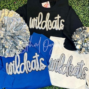 May include: Black, blue, and white sweatshirts with the word "wildcats" in sparkly silver lettering. The sweatshirts are displayed with silver pom-poms. The brand name "Stitched Originals" is also visible.