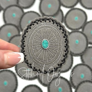 May include: A gray felt circle with a black embroidered border and a silver wheel design. The center of the wheel has a turquoise blue gemstone.