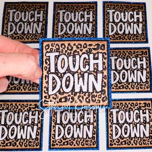 May include: Square embroidered patches with the words "TOUCH DOWN" in white, outlined in black, on a leopard print background. The patches have a blue glitter border. The patches are for sports fans.
