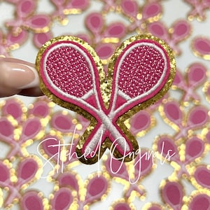 May include: Pink and gold glitter iron-on patch of two crossed tennis rackets. The patch is perfect for adding a sporty touch to clothing, bags, or other accessories.