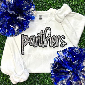 May include: White sweatshirt with the word "Panthers" in black with silver glitter lettering. The sweatshirt is surrounded by blue and silver pom-poms.