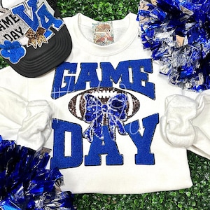 May include: White sweatshirt with blue glitter lettering that says "GAME DAY" with a football and a blue bow with silver glitter.