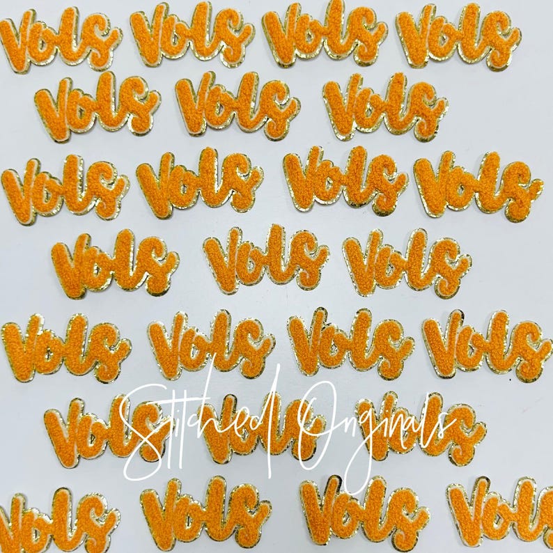 May include: A collection of twenty-two orange felt patches with gold trim, each featuring the word "Vols" in a stylized script font. The patches are arranged in a random pattern on a white surface.