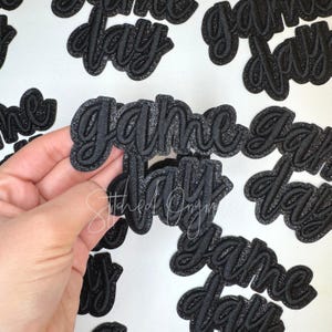 May include: Black glittery "Game Day" patches in a cursive font. The patches are embroidered with a black thread and have a glittery texture. The patches are scattered on a white surface.