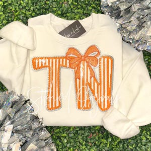 May include: Cream-colored sweatshirt with orange and silver sequined "TN" letters and a bow design. The letters have vertical stripes. The sleeves are visible on either side of the sweatshirt. The background is green.