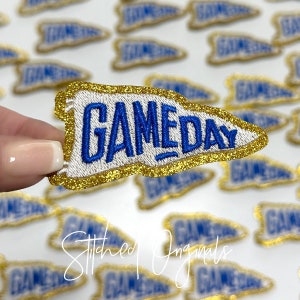 May include: A gold glitter iron-on patch with a white felt pennant shape and blue embroidered text that reads "GAMEDAY".