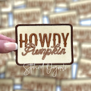 May include: Rectangular patch with the words "HOWDY Pumpkin" embroidered in brown thread on a cream-colored background. The patch has a brown border and the words "Stitched Originals" in a cursive font.