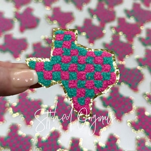 May include: A pink and teal checkered Texas shaped sticker with a gold glitter outline. The sticker is on a white background.