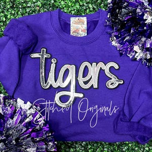 May include: Purple sweatshirt with the word "tigers" in silver glitter lettering. The sweatshirt has a crew neck and long sleeves. The word "tigers" is outlined in black. The text "Stitched Originals" is written in white cursive below the word "tigers".