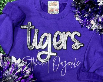 May include: Purple sweatshirt with the word "tigers" in silver glitter lettering. The sweatshirt has a crew neck and long sleeves. The word "tigers" is outlined in black. The text "Stitched Originals" is written in white cursive below the word "tigers".