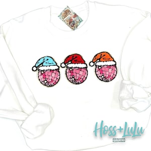 May include: White sweatshirt with three pink disco ball ornaments wearing Santa hats in red, orange, and blue. The hats have gold trim. The sweatshirt has the text "Hoss + Lylu Stitched" in black.