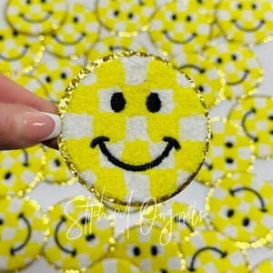 May include: A yellow and white checkered smiley face patch with a gold glitter trim. The patch is made of fuzzy fabric and has a black embroidered smile.