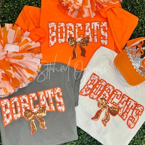 May include: An assortment of orange, white, and gray apparel with the word "BOBCATS" in a decorative font. The apparel includes t-shirts and a baseball cap. Each item has a sequined bow. Pom-poms are also visible.