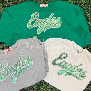 May include: Three crewneck sweatshirts in green, gray, and white, each featuring the word Eagles in cursive script. The lettering is crafted from green glitter material with a silver glitter outline. Each garment has a small interior brand label.