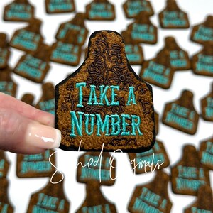May include: Brown leather-like patch with a floral design and the text "Take a Number" in turquoise blue embroidery. The patch is shaped like a cow tag.