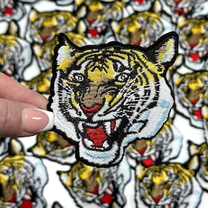 May include: Close-up of a white and black embroidered tiger face patch with a red mouth and yellow stripes. The patch is surrounded by other tiger face patches.