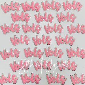 May include: A collection of pink felt iron-on patches with the word "Vols" in a cursive font. The patches have a silver outline.