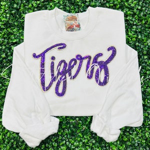 May include: White sweatshirt with the word "Tigers" in large, sparkling purple letters. The word is outlined in gold and has a cursive font. The sleeves are long and cuffed. A small tag is visible near the collar.