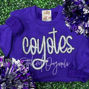 May include: Purple sweatshirt with the word "Coyotes" in silver glitter lettering. The sweatshirt has a crew neck and long sleeves. The word "Stitched Originals" is also visible in silver glitter lettering.