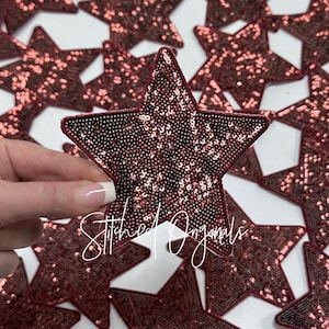 May include: A close-up of a pile of burgundy star-shaped fabric patches with sequins. The patches are arranged in a random pattern and are all the same size. The word "Stitched Originals" is visible in the lower right corner of the image.