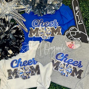 May include: Three sweatshirts in blue, white, and gray, each with the text "Cheer Mom" in sparkling blue and silver embellishments. A pom-pom and a foam finger are also visible.