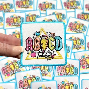 May include: A colorful embroidered iron-on patch with the letters ABCD in a rainbow color scheme. The letters are surrounded by a triangle with a lightning bolt, an apple, a pencil, and a book. The text "The Teacher Tour" is below the letters.