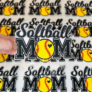 May include: Embroidered patch with the words "Softball MOM" in black and white. A yellow softball with a black heart is in the center. The patch is surrounded by other similar patches.