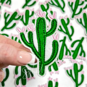 May include: A green cactus with pink flowers embroidered on a white fabric patch. The cactus is a popular motif for crafting and decorating.