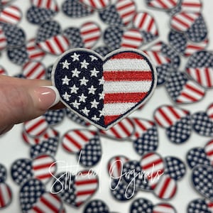 May include: A close-up of a hand holding a heart-shaped embroidered patch with the American flag design. The patch is surrounded by other heart-shaped patches with the American flag design.