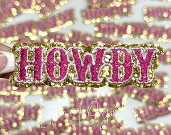 May include: Pink and gold glitter iron-on patch with the word "HOWDY" in a bold, cursive font.