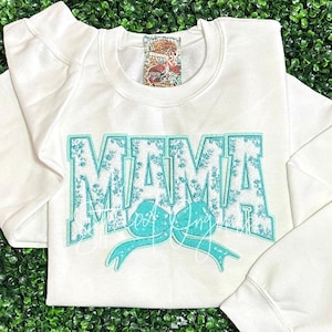 May include: White sweatshirt with a turquoise blue "MAMA" graphic with a floral pattern and a turquoise blue bow.