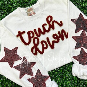 May include: White sweatshirt with the words "touch down" in maroon felt lettering with gold outlining. The sweatshirt has maroon sequined stars on the sleeves.