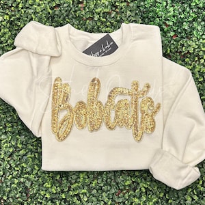 May include: Cream-colored sweatshirt with the word "Bobcats" in gold sequins. The long-sleeved shirt has a crew neck and a tag that reads "Hugs + Lulu". The shirt is folded on a green surface.