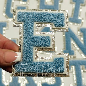 May include: A light blue, fuzzy letter "E" patch with a silver glitter border and white backing. The patch is held in a hand, with other letter patches visible in the background. The patch is likely for clothing or crafts.